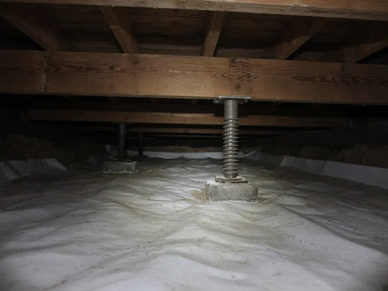 Crawl space with an adjustable steel support post under a wood beam and a white plastic vapor barrier on the ground